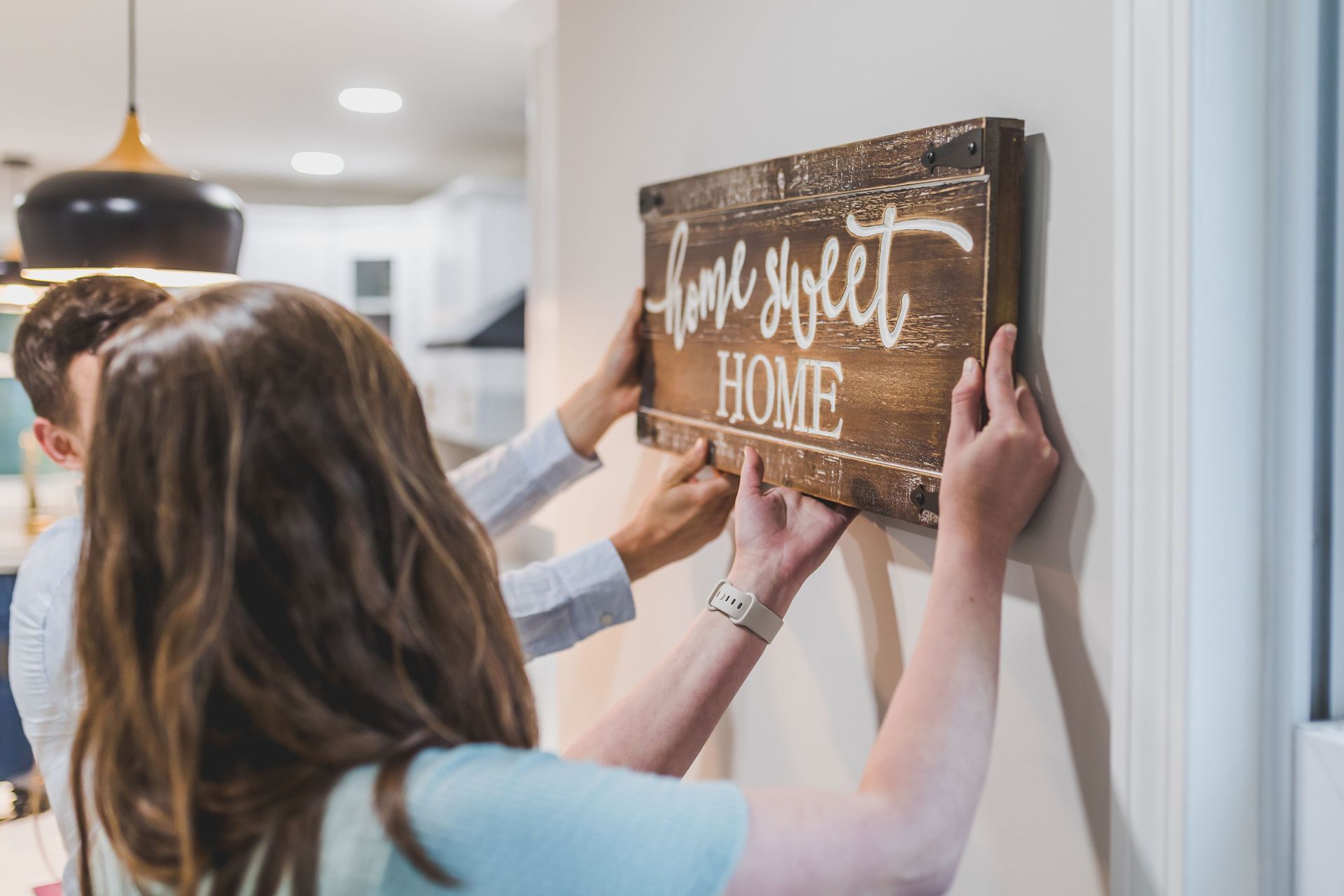 Two people hang a rustic “love sweet home” sign in a bright hallway.
