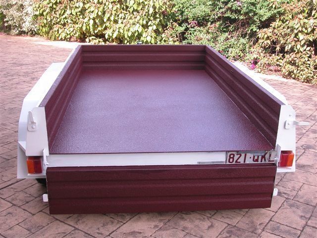 Rear view of an empty maroon trailer with white trim, parked on a paved surface, against a background of foliage.