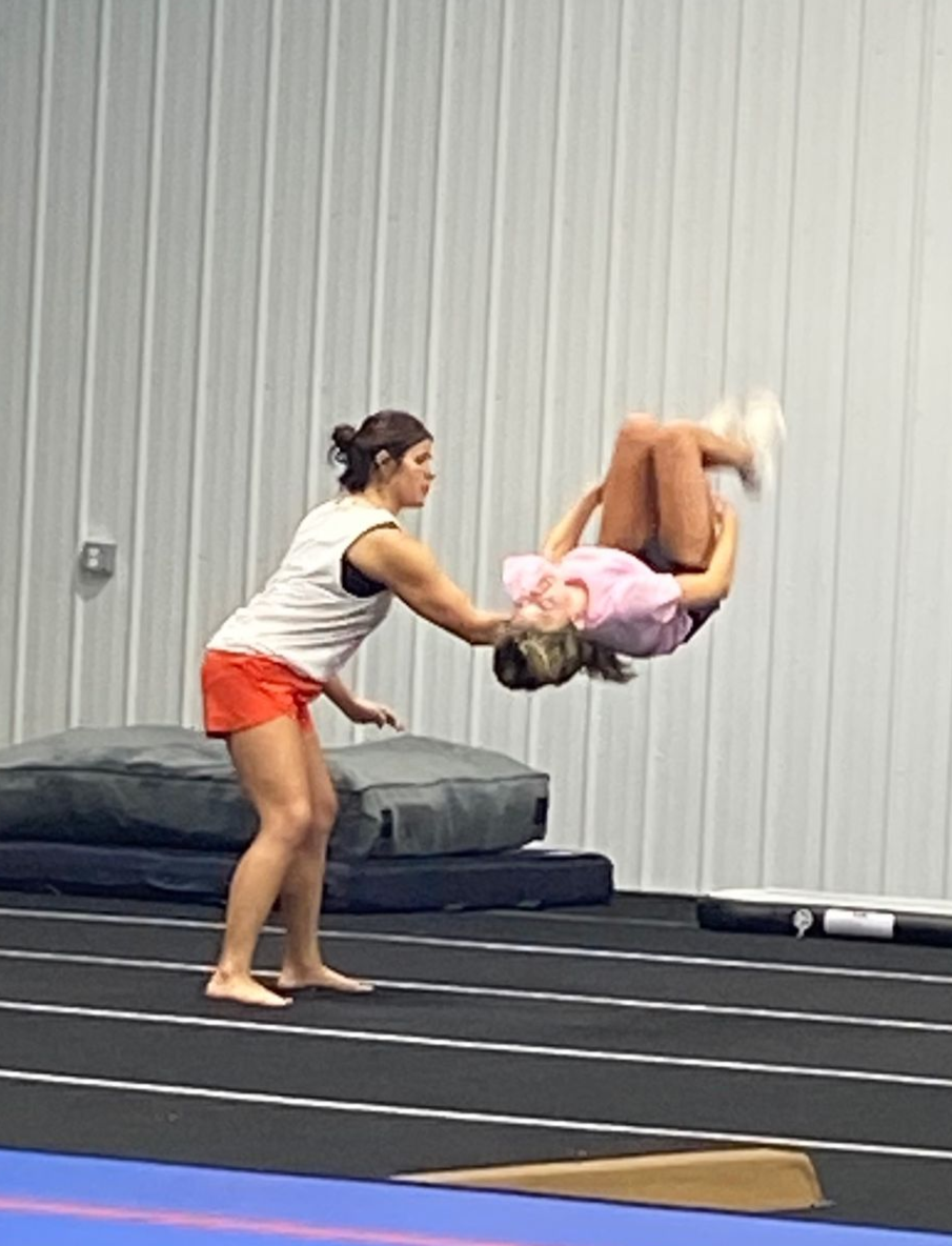 Cheerleader in black and red uniform doing a back tuck in a gymnasium. Her ponytail has a red bow.