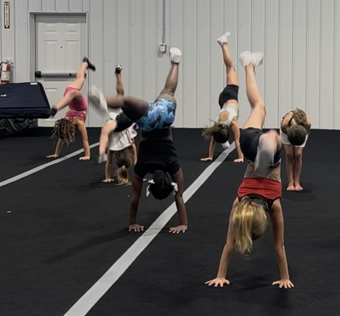 Young girls practicing handstands on a black mat in a gym. Some have white shoes, and the gym has a white door and lines.