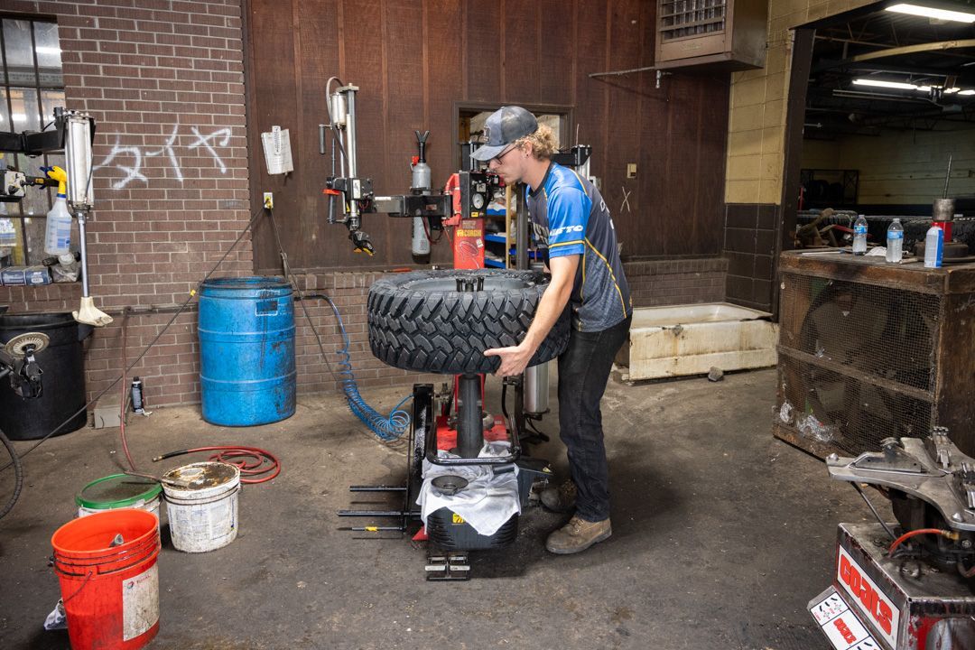 Mechanic working on a car tire in a cluttered auto shop with tools and buckets nearby