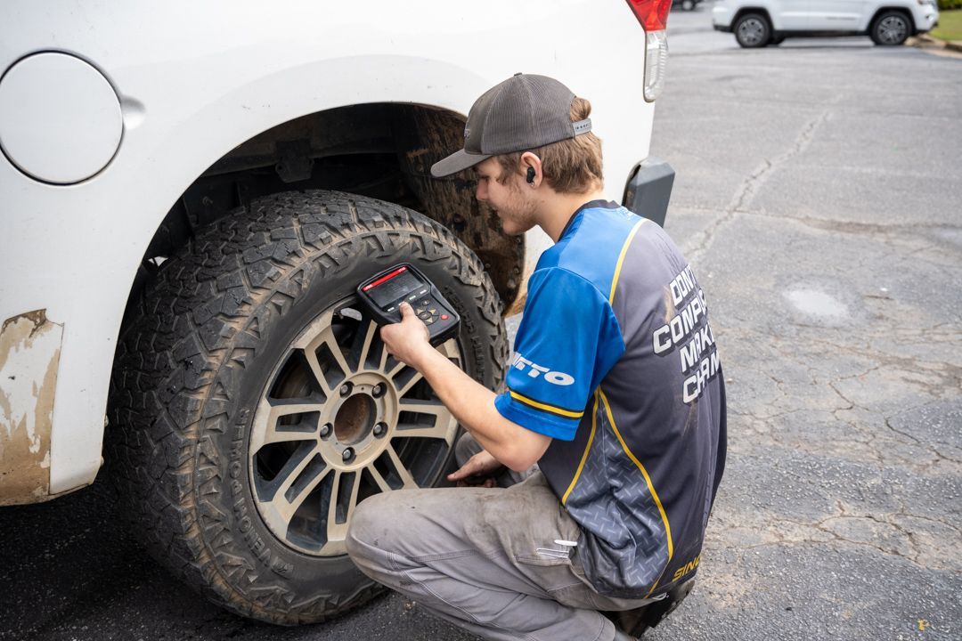 Mechanic kneeling by a white car, checking a muddy tire with a diagnostic device