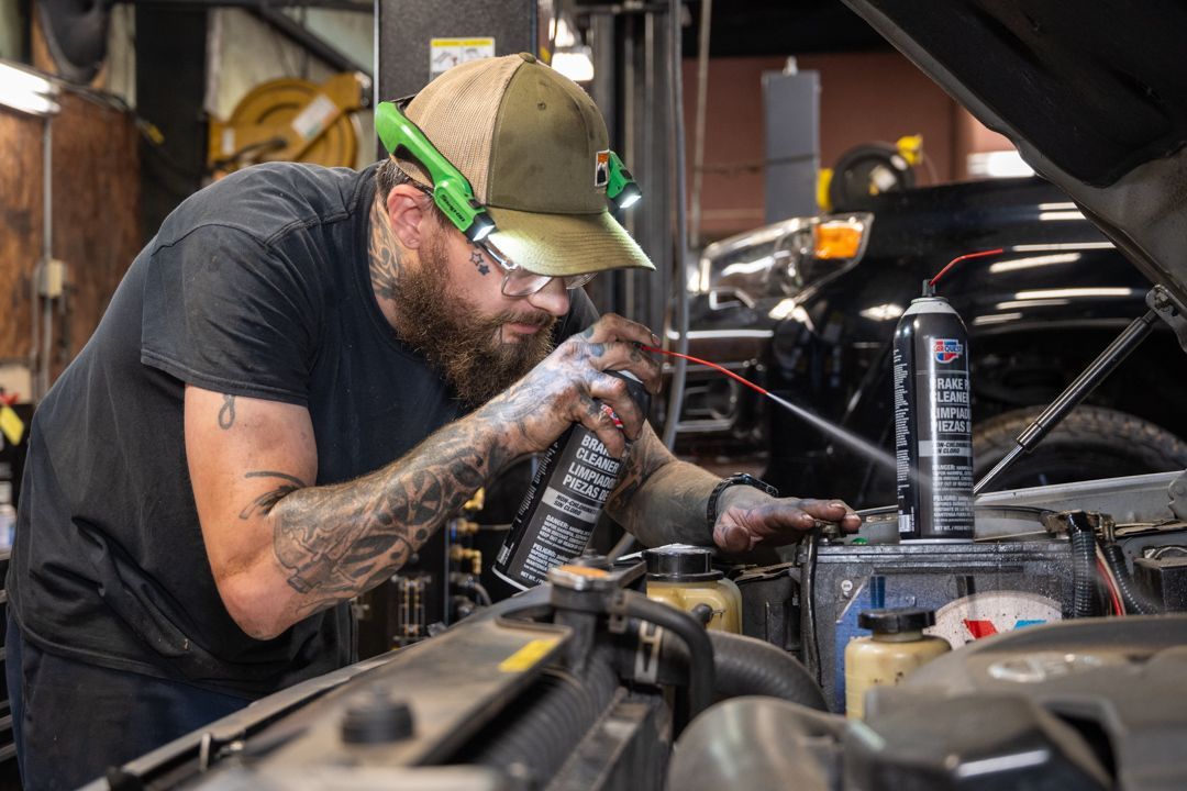 Mechanic inspecting car engine in a garage, wearing gloves and a cap, with tools and parts nearby