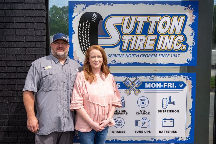 Two people standing beside a Sutton Tire Inc. business sign outdoors.