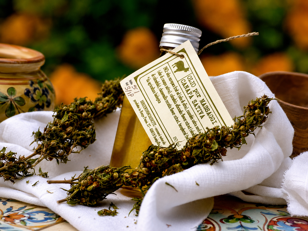 Una piccola bottiglia di vetro e dei rametti di erbe aromatiche poggiano su un panno bianco, con un vaso decorativo sullo sfondo.