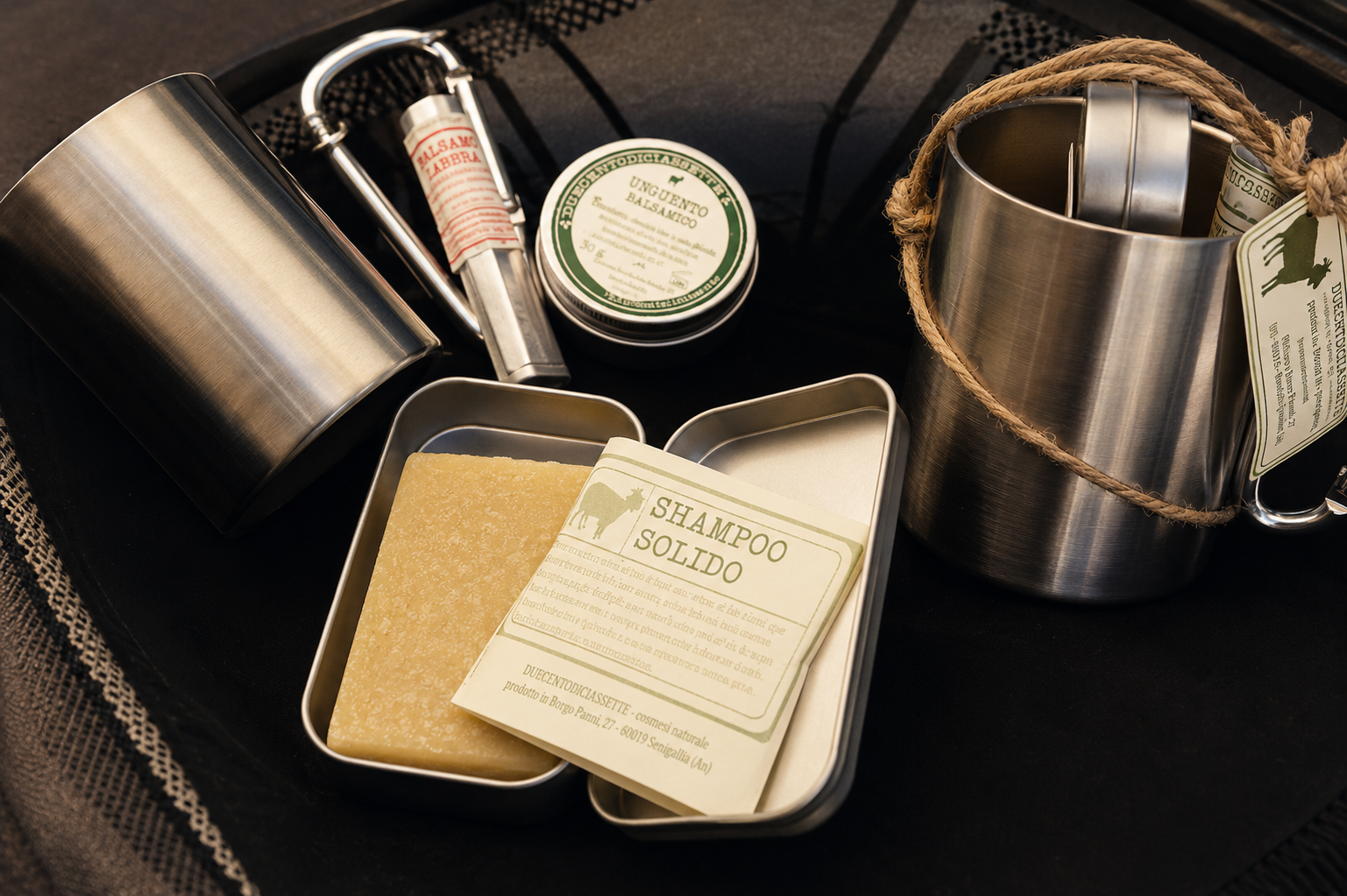 Kit da viaggio a righe bianche e nere con contenitori in latta, portasapone e biglietto piegato su una superficie scura.