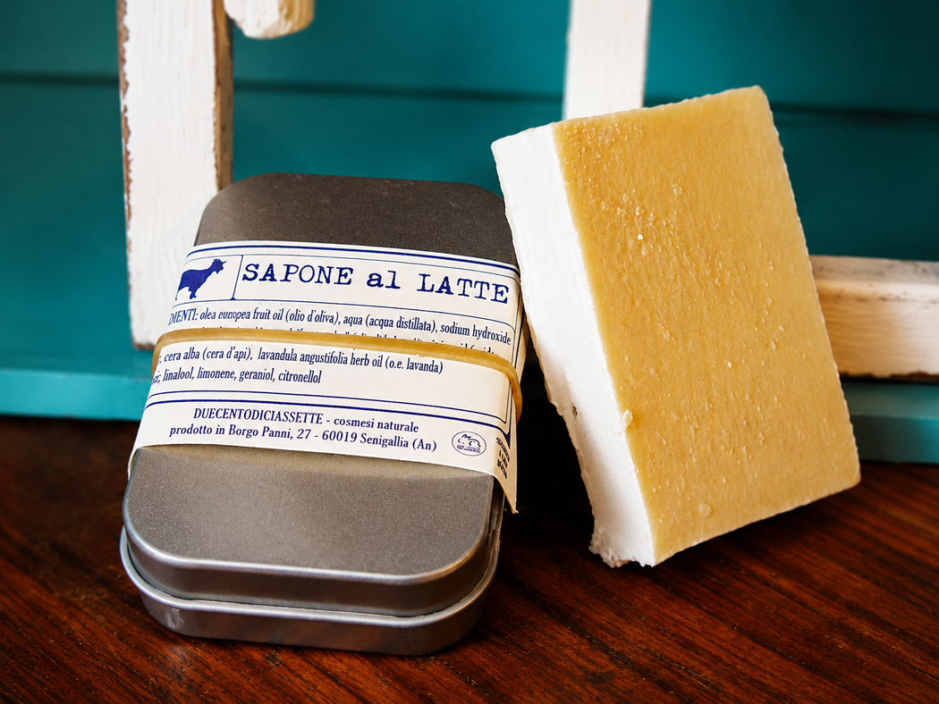 Due saponette in una scatola di metallo, una incartata e l'altra no, su un tavolo di legno.