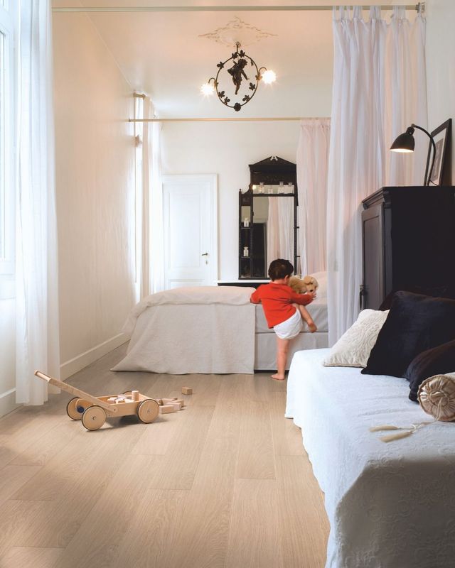 Child in red shirt climbing onto a white bed, light wood floor, white curtains and walls, black furniture.