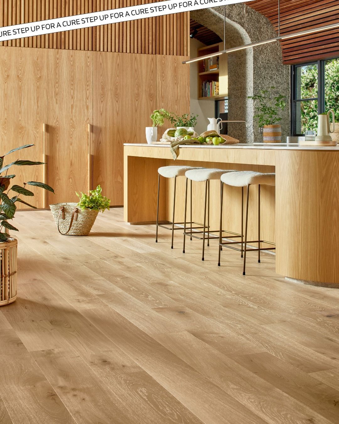 Wooden kitchen with wood flooring, island, and stools. Natural light, plants, and a light-colored countertop.