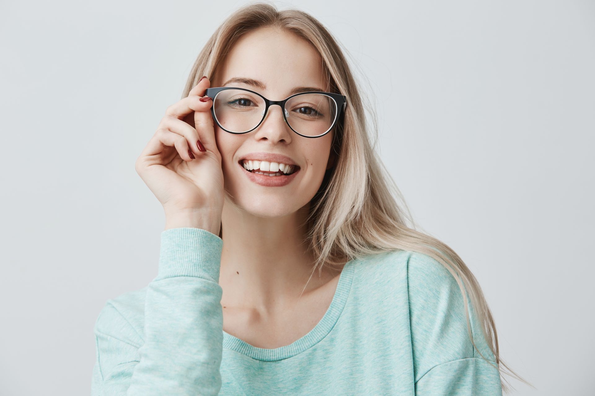 Smiling person in a light blue sweater adjusting black glasses on a plain white background