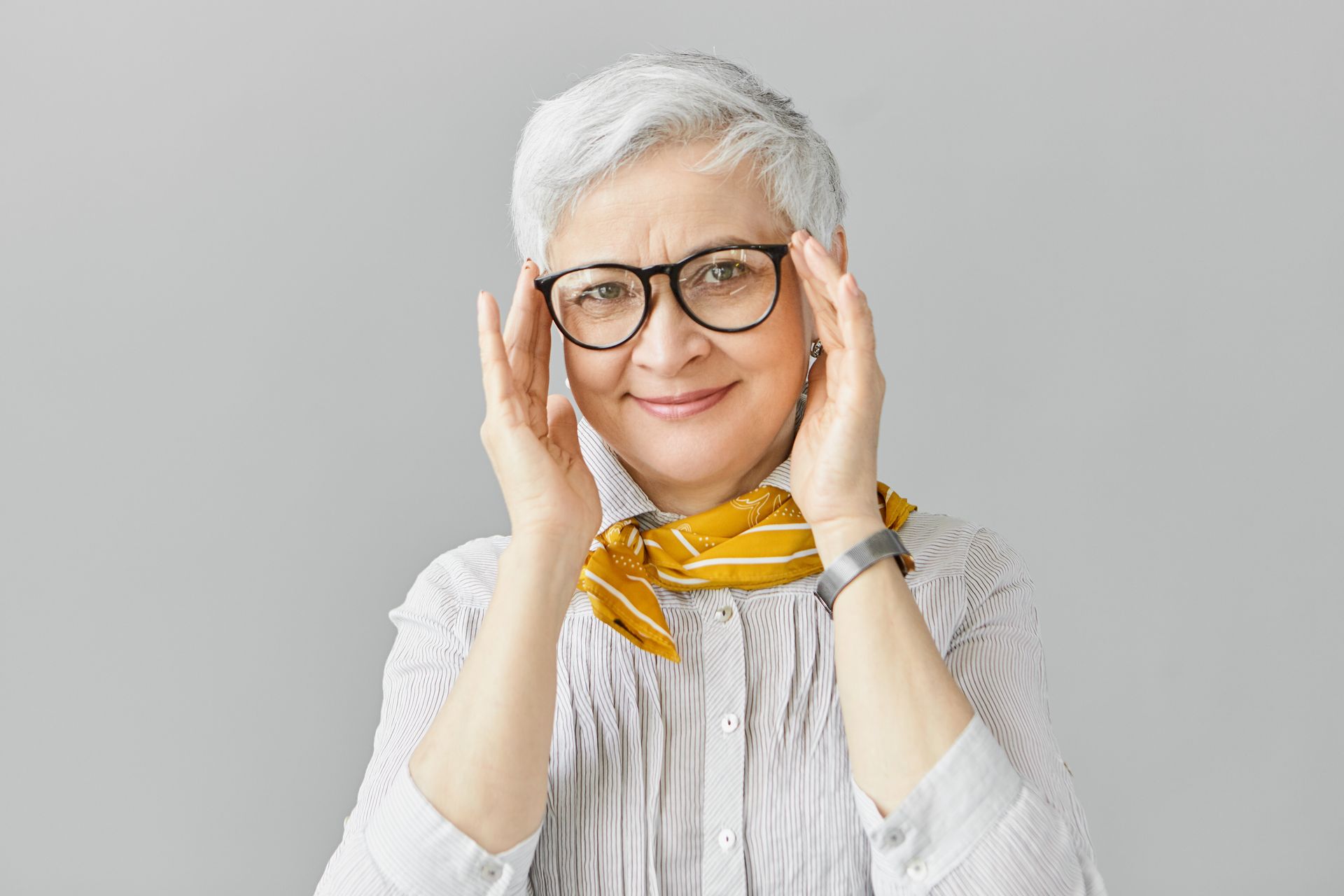 Smiling woman with short white hair, glasses, and yellow scarf touching her face against a gray background