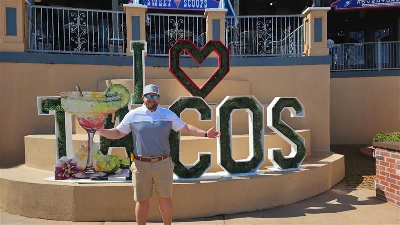Person posing with arms out in front of a colorful “I ♥ TACOS” sign outdoors