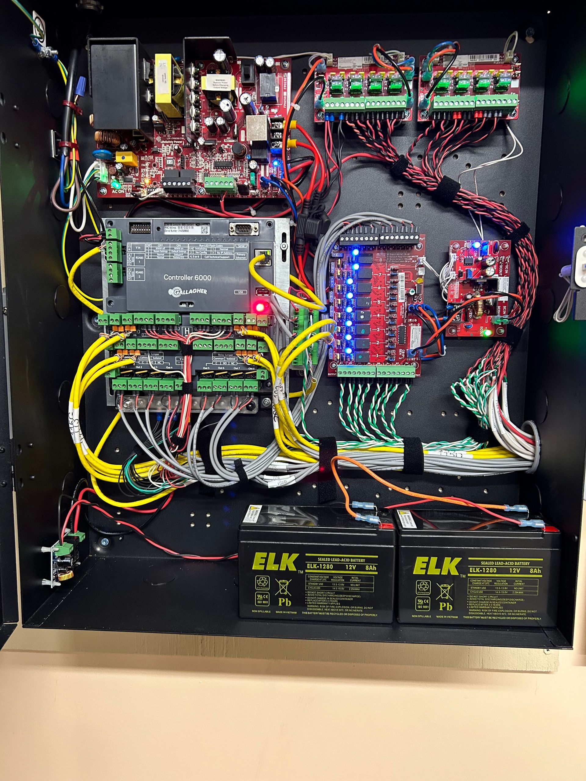 Open electronics control panel with dense wiring, circuit boards, relays, and two black batteries.