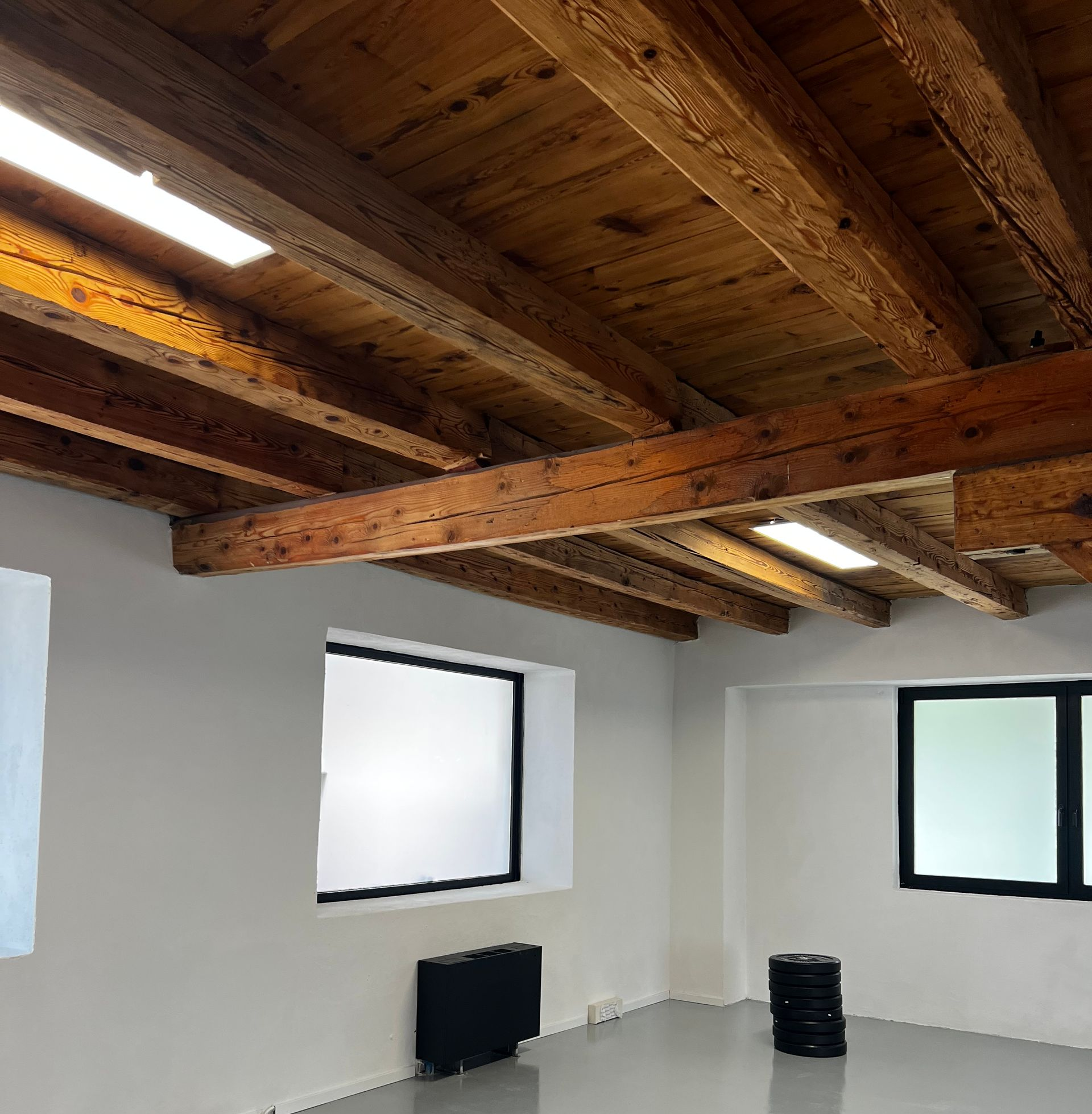 Una sala fitness con travi in legno a vista, pareti bianche e pile di dischi per bilanciere neri su un pavimento grigio.