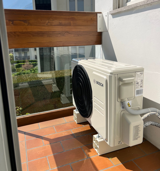 Un'unità esterna di un condizionatore d'aria Bosch di colore beige è installata su un balcone con piastrelle in terracotta e ringhiera in legno e vetro.