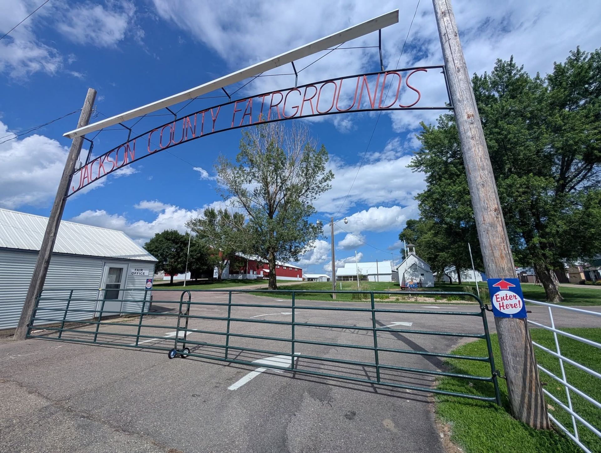 A metal archway sign reading 'McLean County Fairgrounds' over a gated entrance to a grassy fairground under a blue sky.