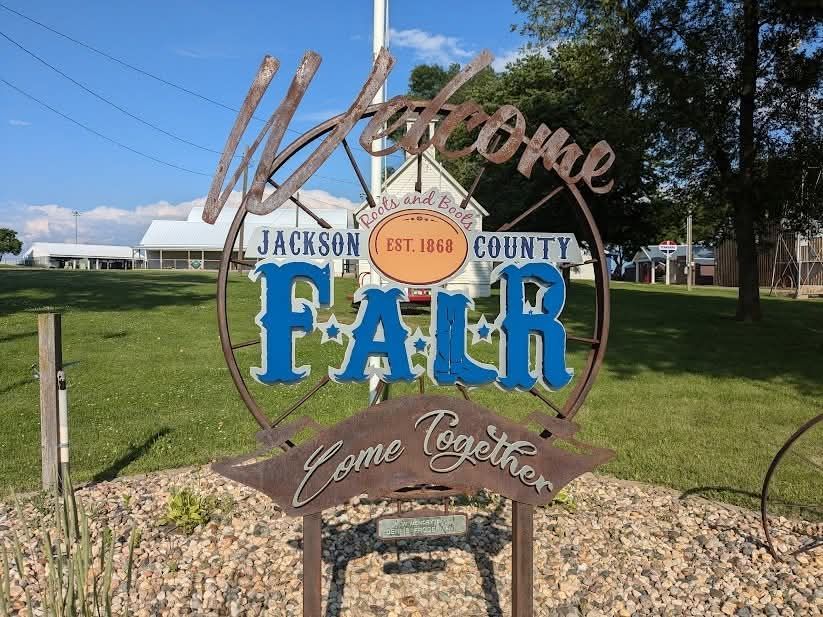 A metal Jackson County Fair welcome sign with blue lettering on a green fairground, with buildings and trees behind.