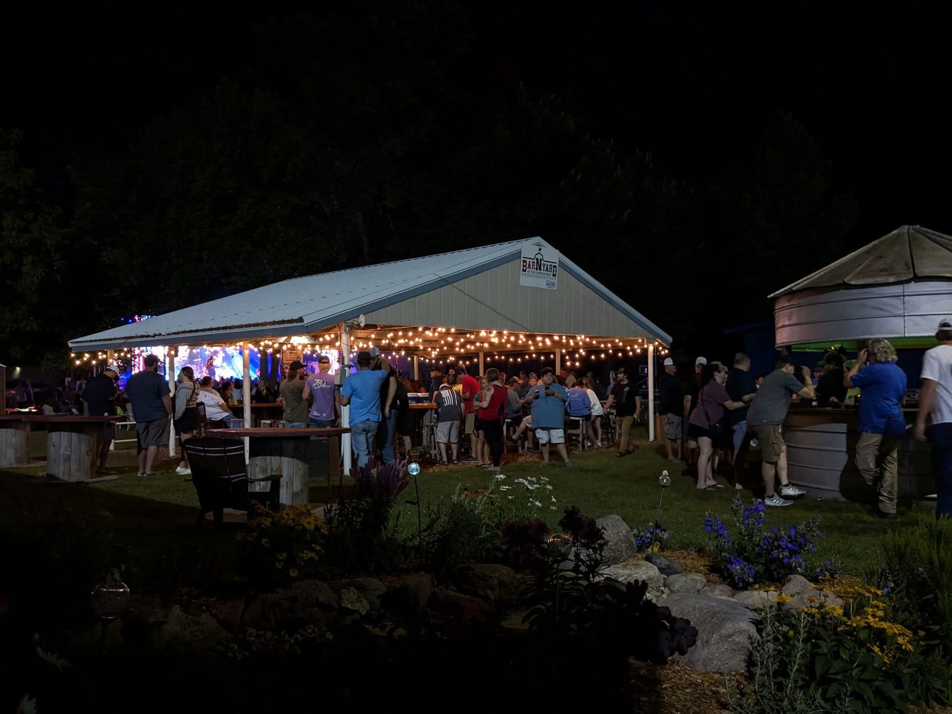 A brightly lit pavilion with string lights hosts a gathering of people at night, with a bar area visible on the right.