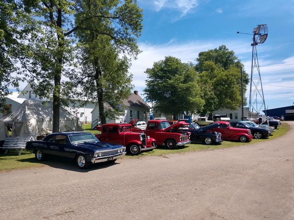 A line of classic vintage cars parked on a gravel lot near trees, white farm buildings, and a tall metal windmill.