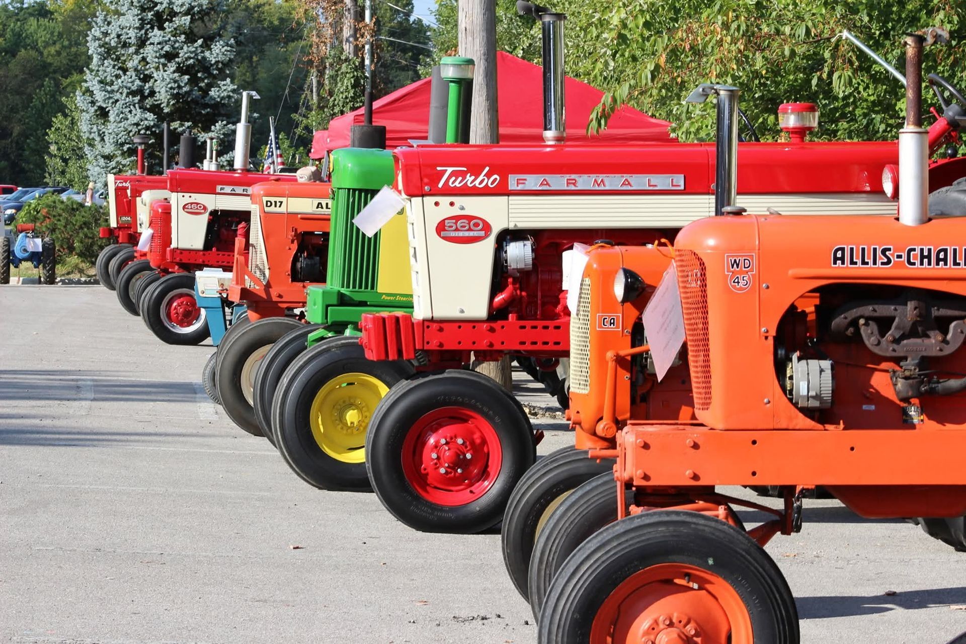 A row of vintage tractors in various colors, including red, orange, and green, parked outdoors in a sunny lot.