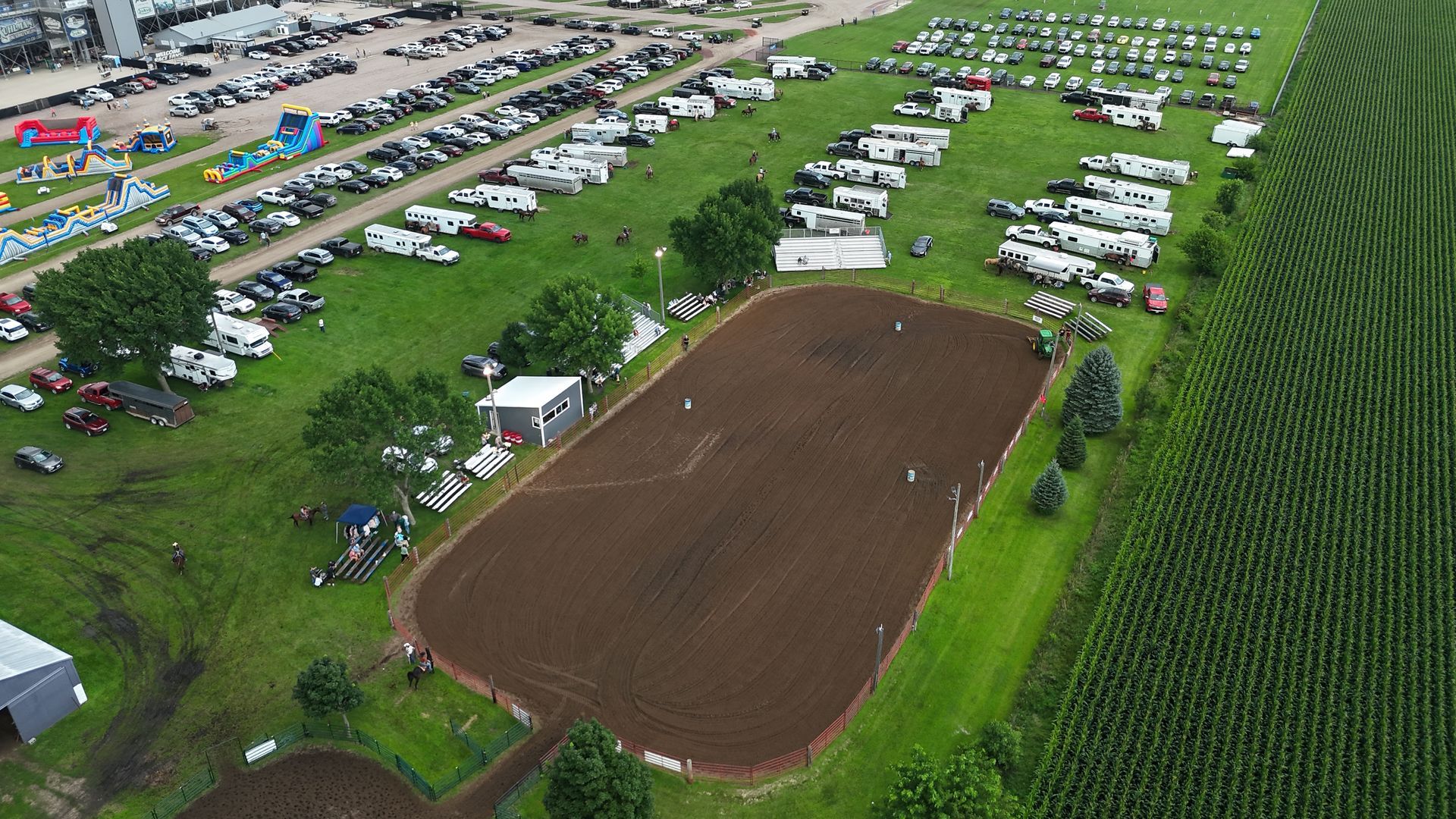 An aerial view shows an equestrian arena surrounded by parked horse trailers, trucks, and a large field of crops.