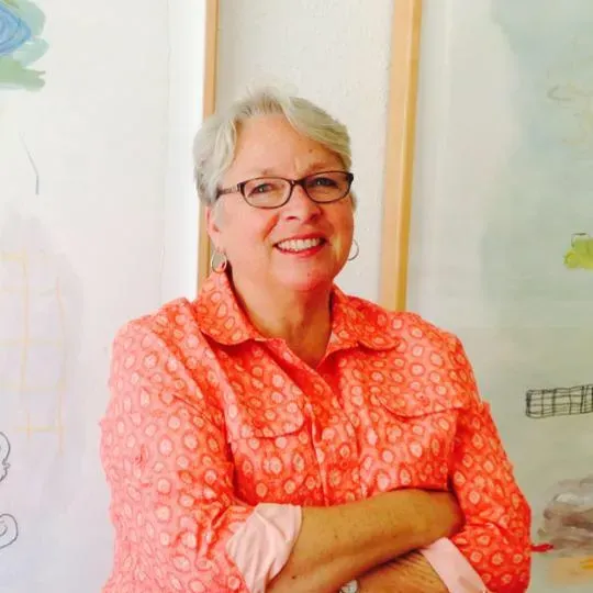 Smiling woman in glasses and a coral patterned shirt, standing with arms crossed against a light wall.