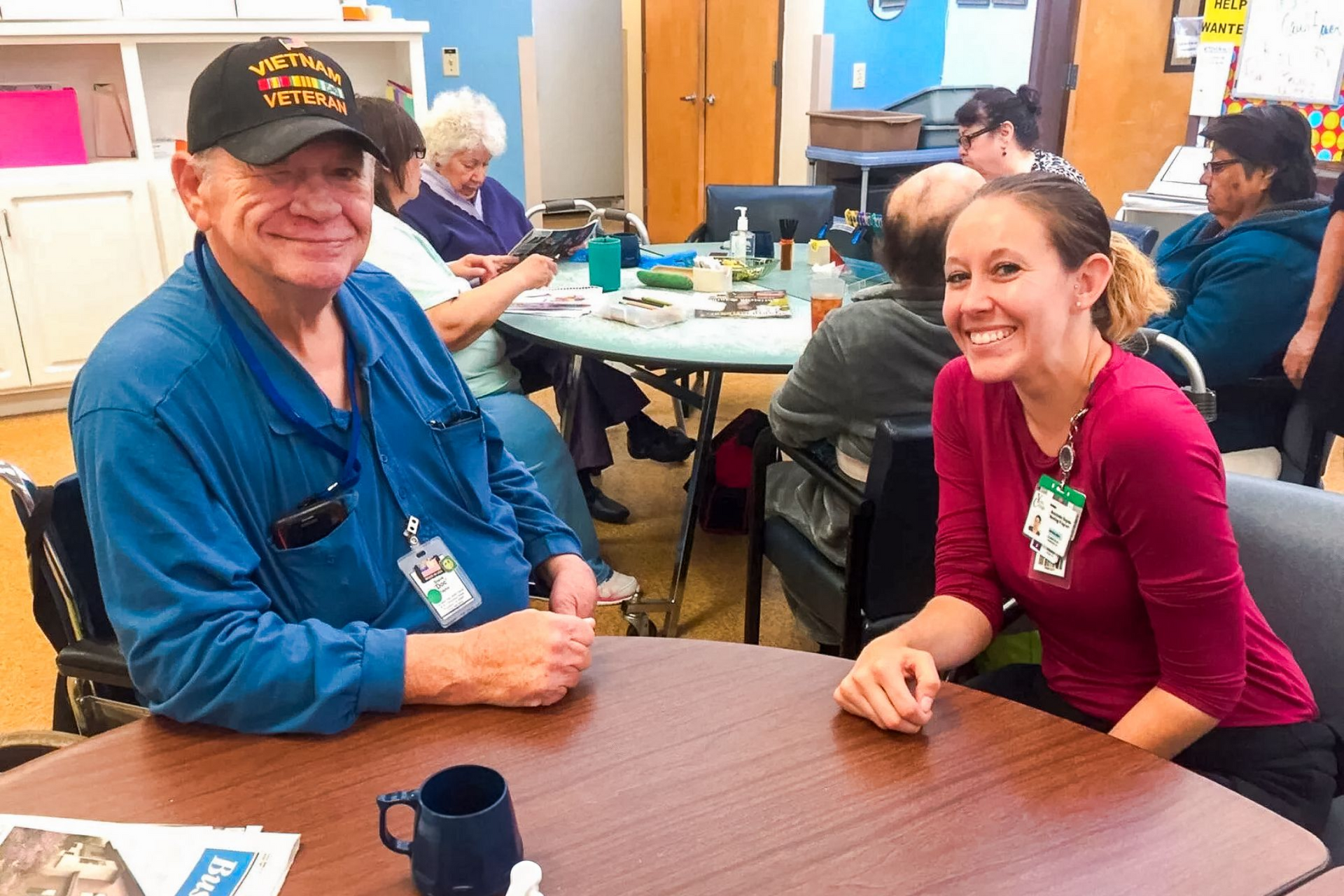 Two people smiling at a table in a community room, with others seated and chatting in the background.