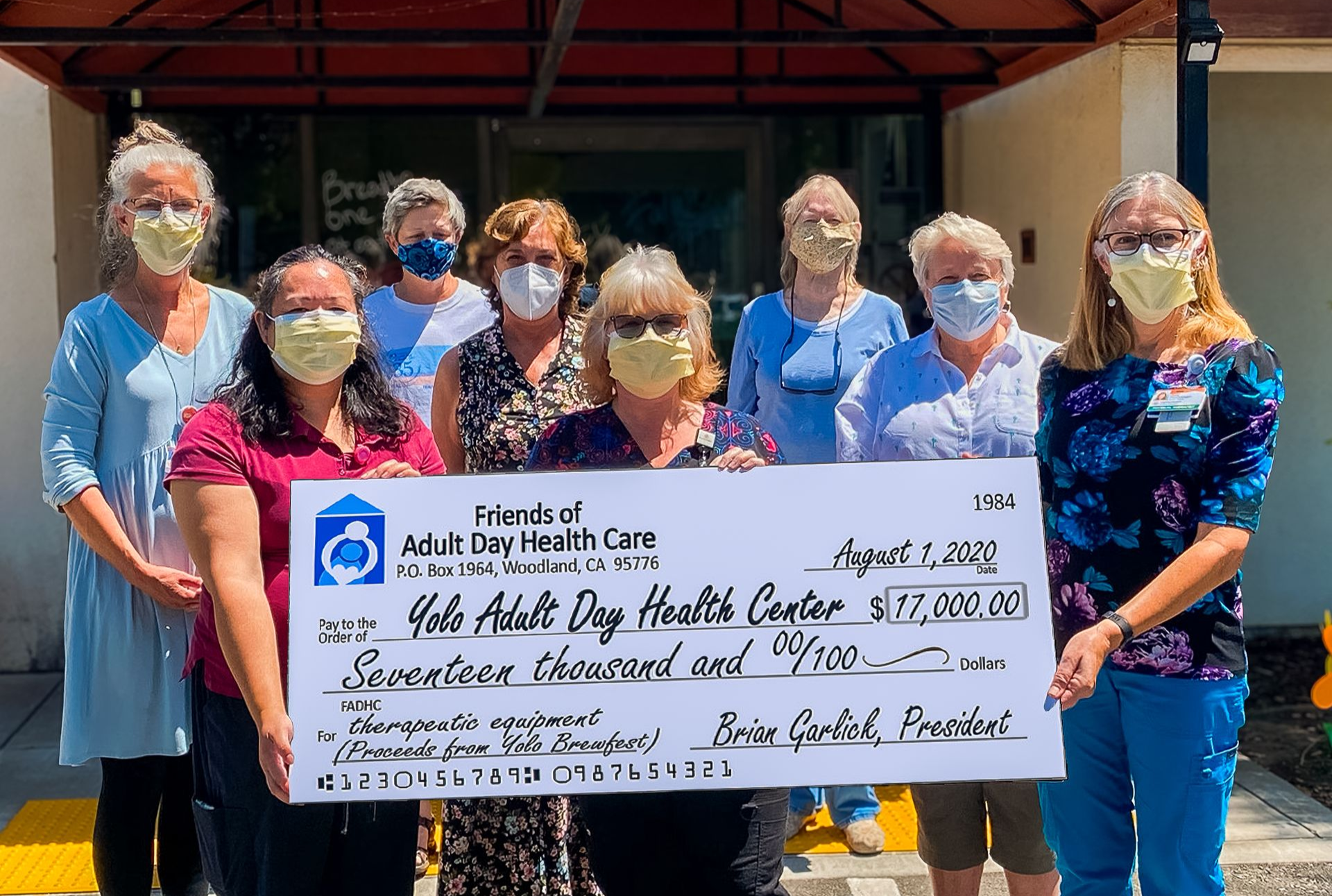 Group holding a large check in front of a building, most wearing face masks and blue scrubs.