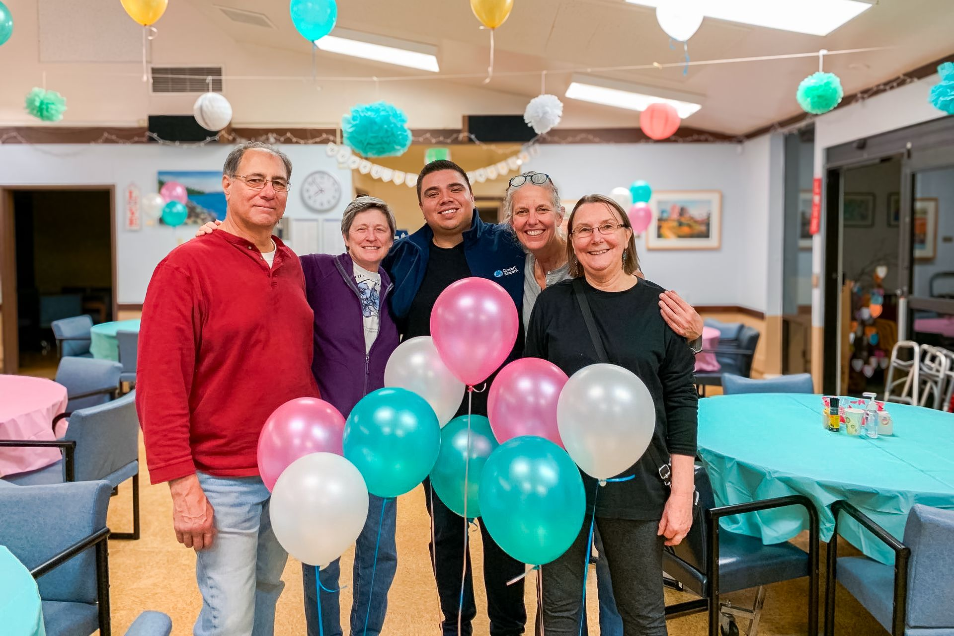 Five people holding colorful balloons in a decorated indoor room, smiling at a celebration