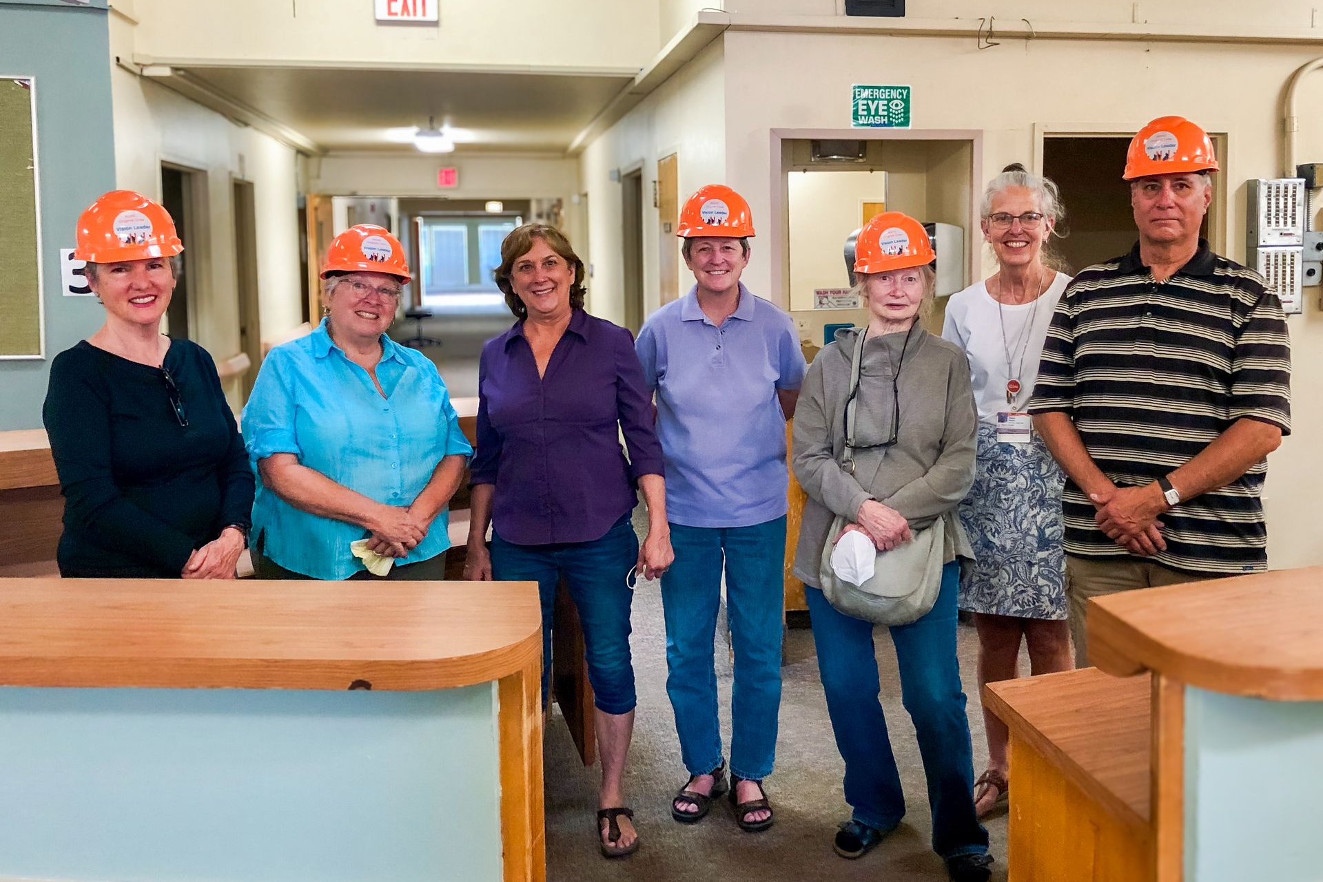 Six people wearing orange hard hats stand in a lobby, posing behind wooden counters.