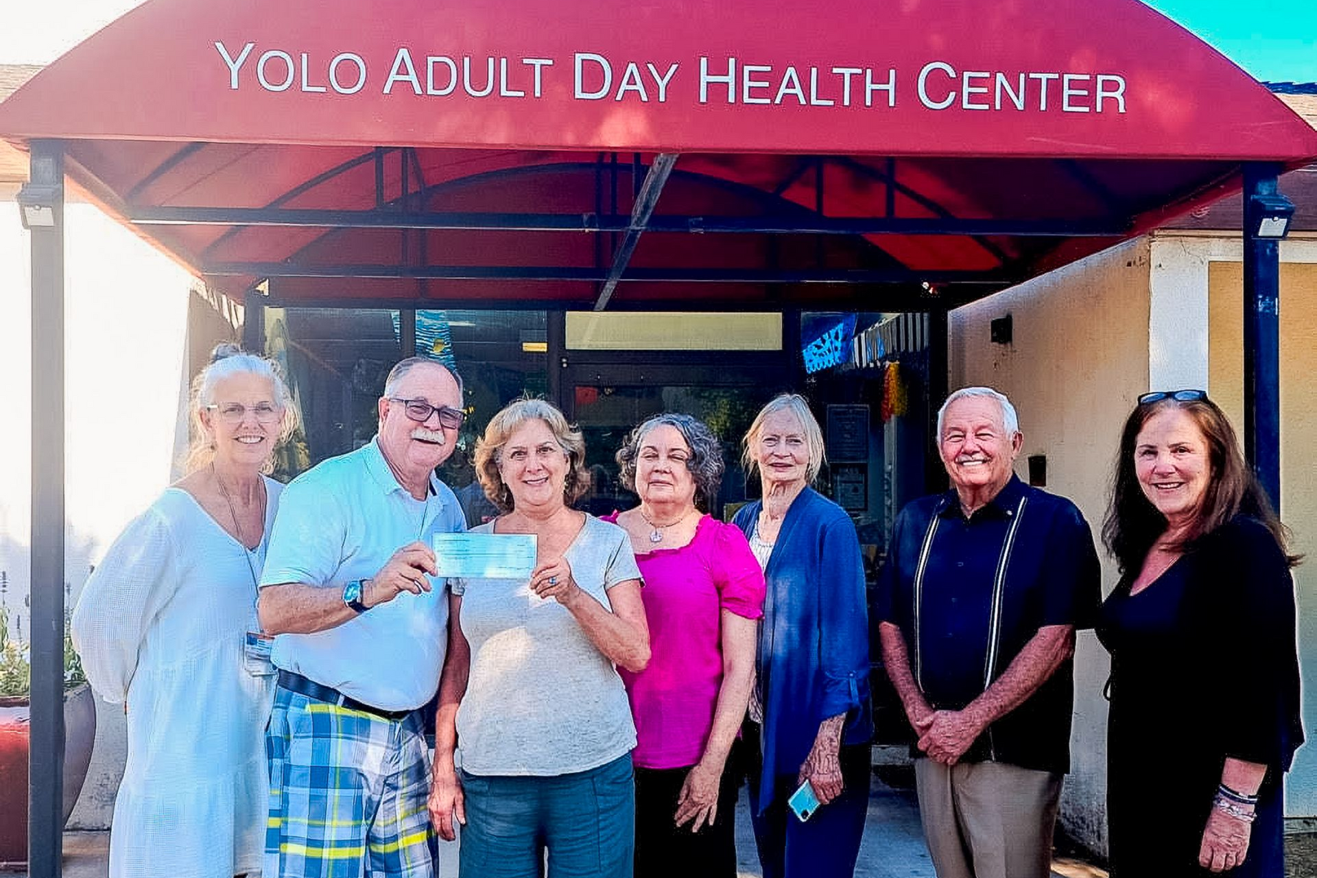 Group of people standing outside Yolo Adult Day Health Center under a red awning, smiling at the camera