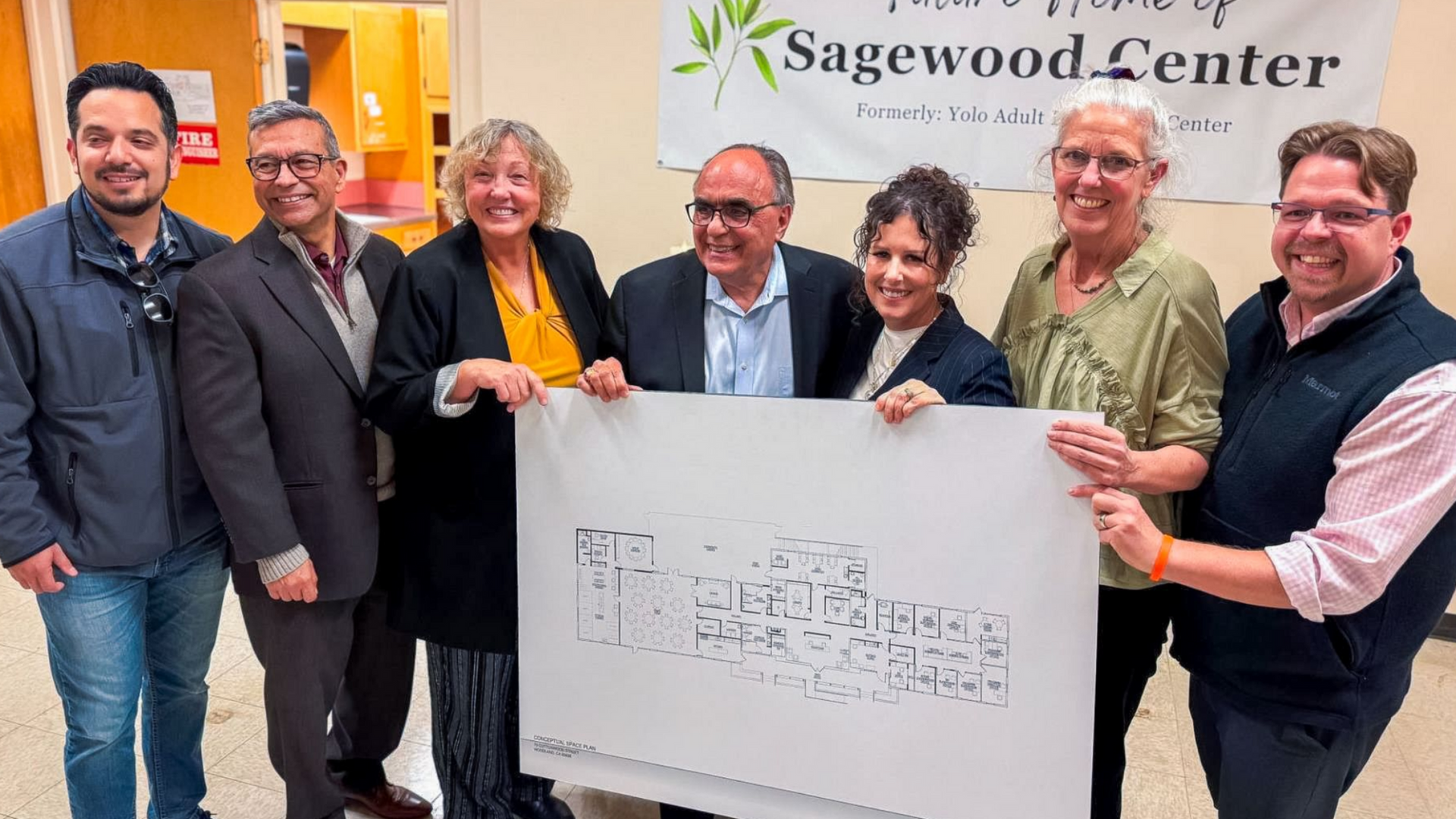 Seven people holding a whiteboard in front of a Sagemod Center sign, smiling indoors