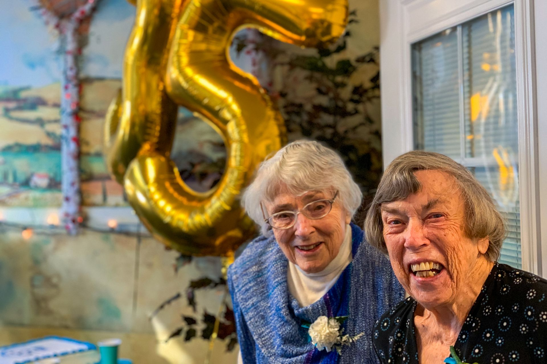 Two smiling people indoors beside a large gold balloon in a festive room