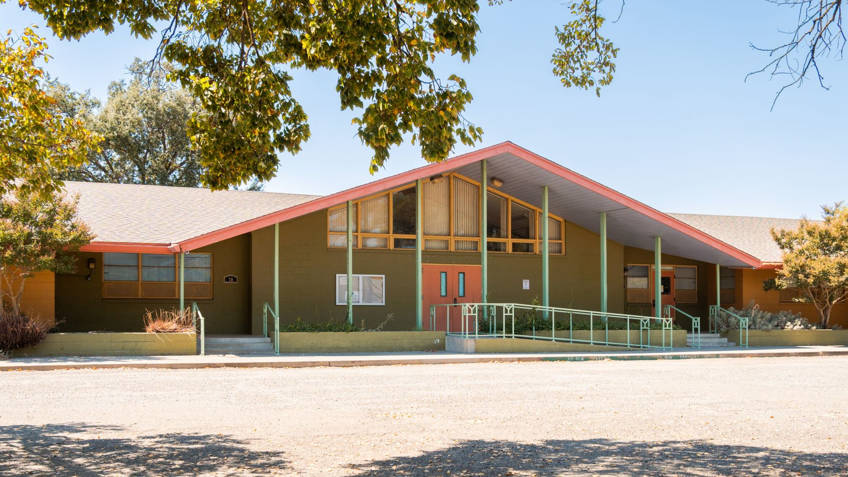Single-story school building with red roof, green walls, and covered entrance under trees