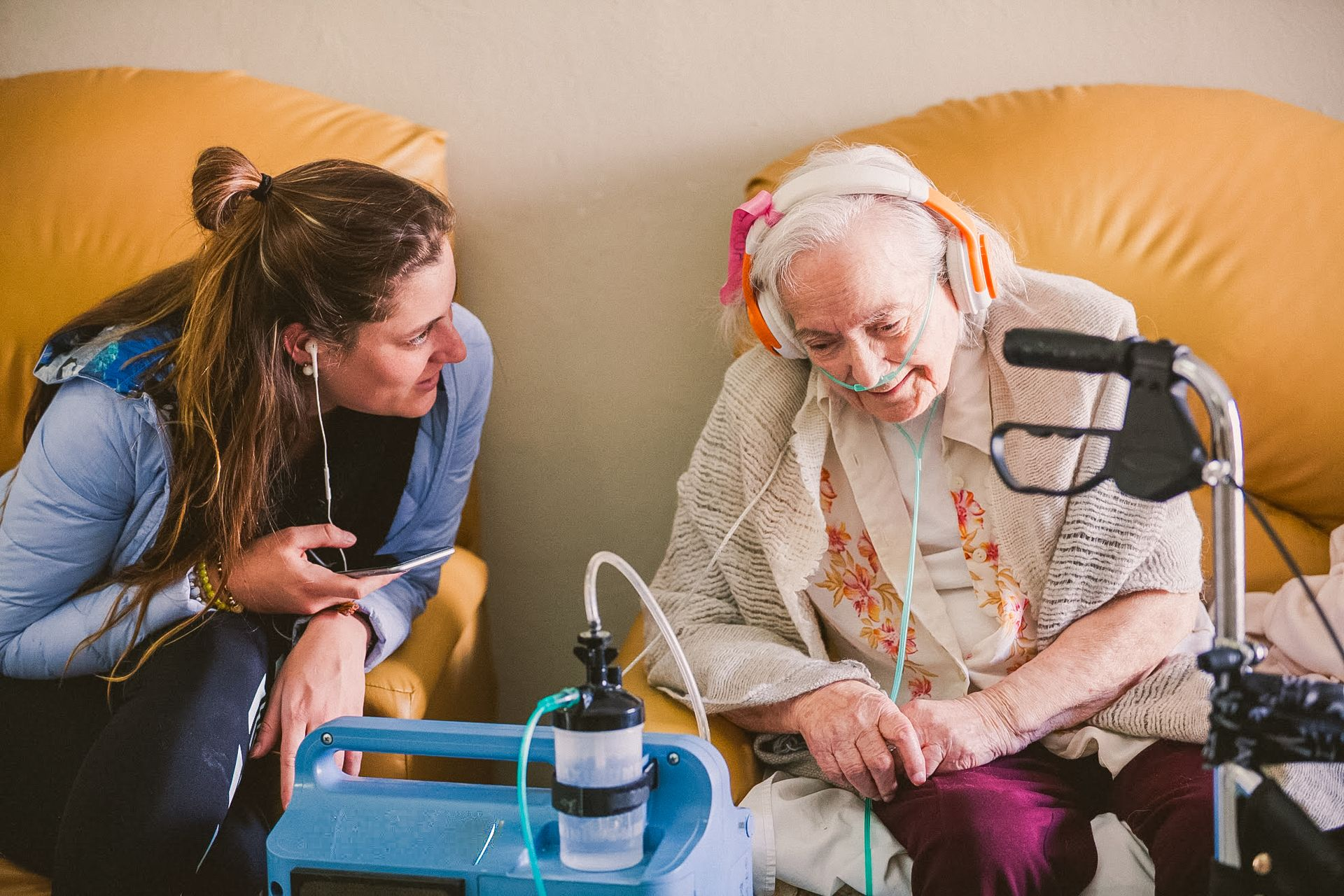 Woman talks with elderly person wearing headset on a couch, beside a medical device and oxygen tank.