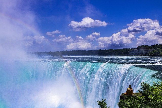 A man is sitting on the edge of niagara falls with a rainbow in the background.