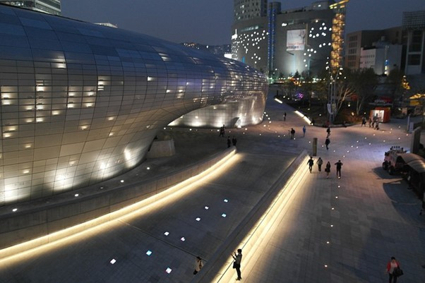 A group of people are walking in front of the Dongdaemun Design plaza, which is lit up at night