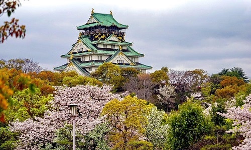 A large castle is surrounded by trees and flowers in a park in Osaka.