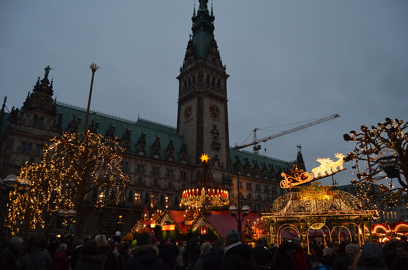 A crowd of people are gathered in front of a hamburg city hall with a christmas market in front of it