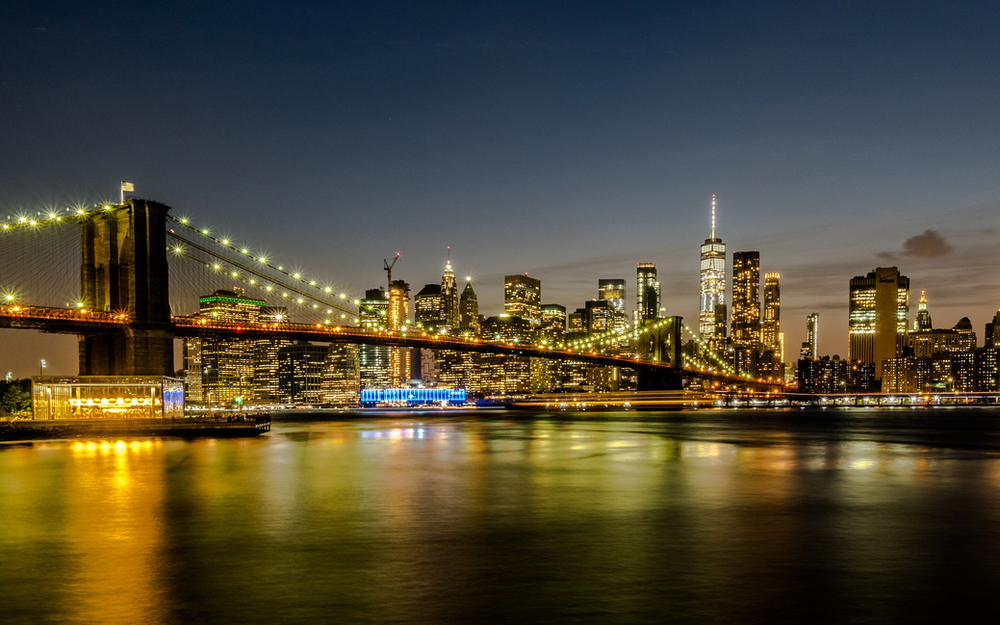A bridge over a body of water with the new york city skyline in the background at night.