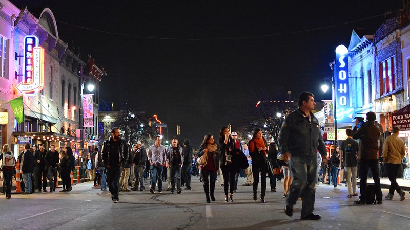 A crowd of people are walking down 6th street in Austin at night.