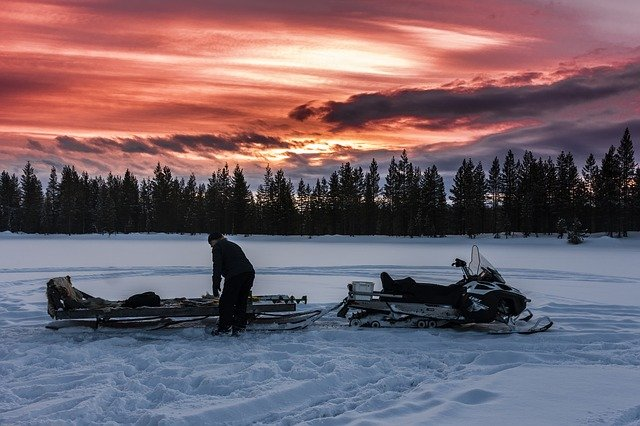 A man is standing next to a snowmobile in the snow at sunset.