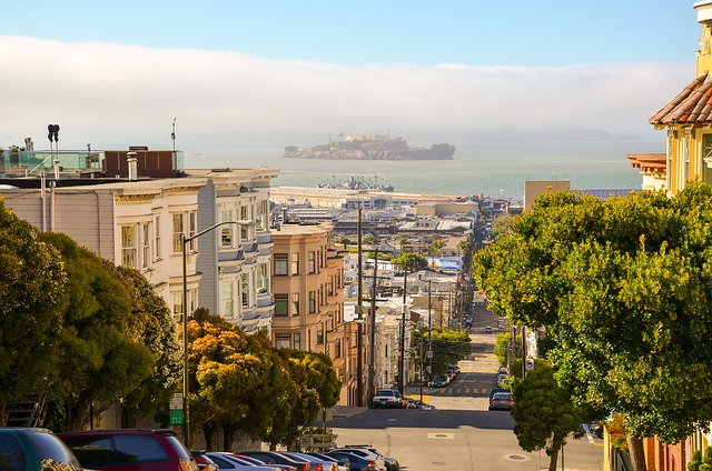 A row of cars are parked on the side of a san francisco street with alcatraz in the background.