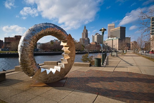 A large metal sculpture is in front of a city skyline on the Providence River Walk