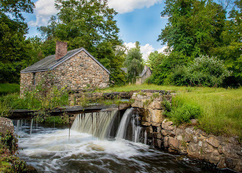 A small waterfall is surrounded by trees and grass in front of a stone house in waterloo village