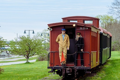 Two men are standing on the back of a train on the maine narrow gauge railroad
