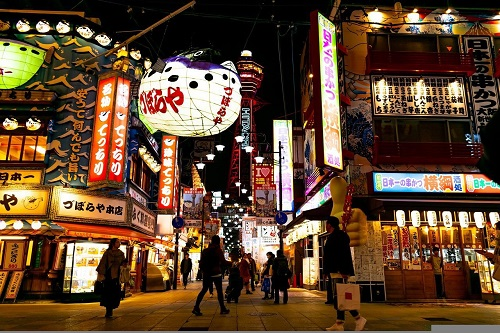 A group of people are walking down a busy street in the Shinsekai district at night
