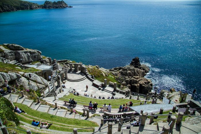 A group of people are sitting in the Minack Theatre amphitheatre overlooking the ocean.