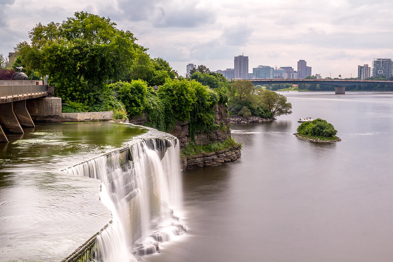 A waterfall in the middle of a river with a city in the background.