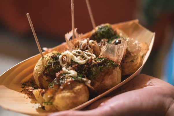 A person is holding a wooden tray of takoyaki with sticks in it.